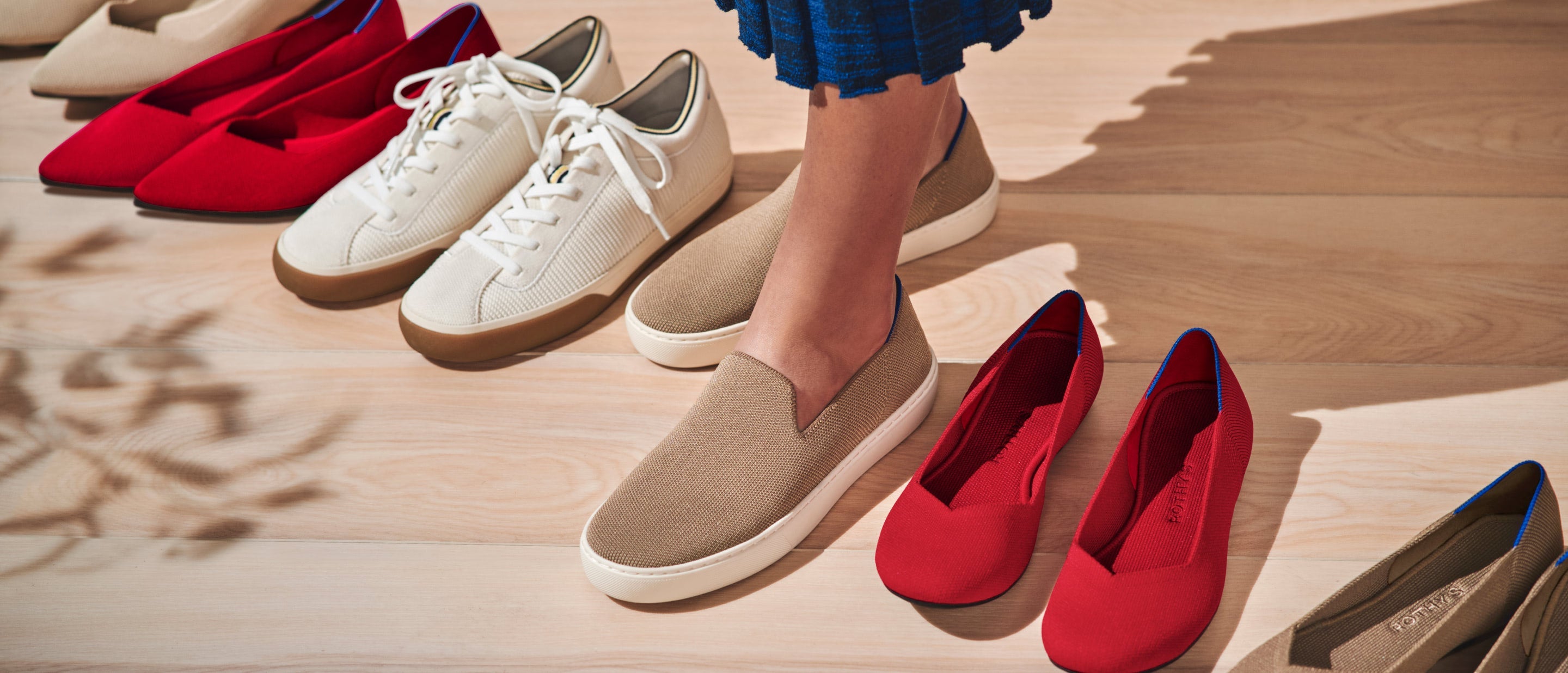 A model wearing The Original Slip On Sneaker in Latte, surrounded by other pairs of Rothy's including The Lace Up Sneaker and The Flat.