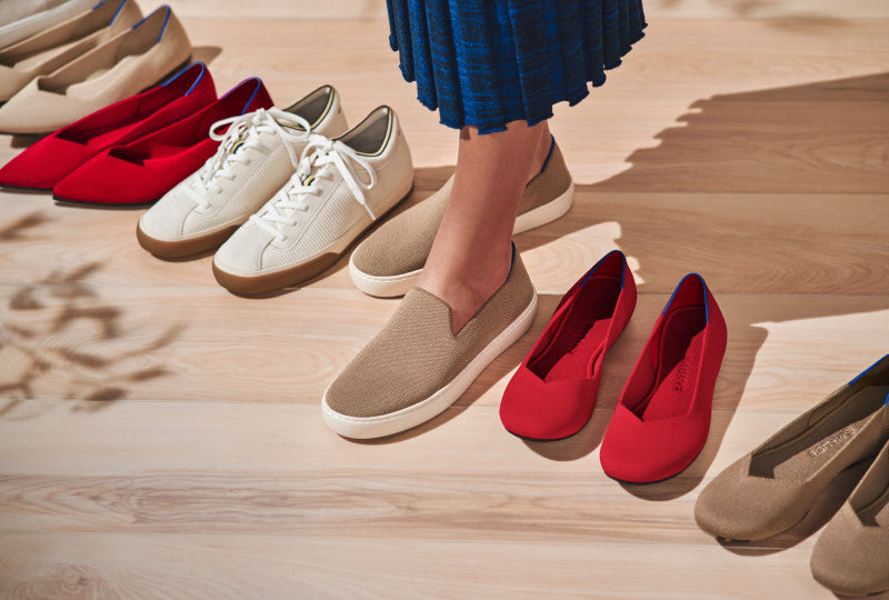 A model wearing The Original Slip On Sneaker in Latte, surrounded by other pairs of Rothy's including The Lace Up Sneaker and The Flat.