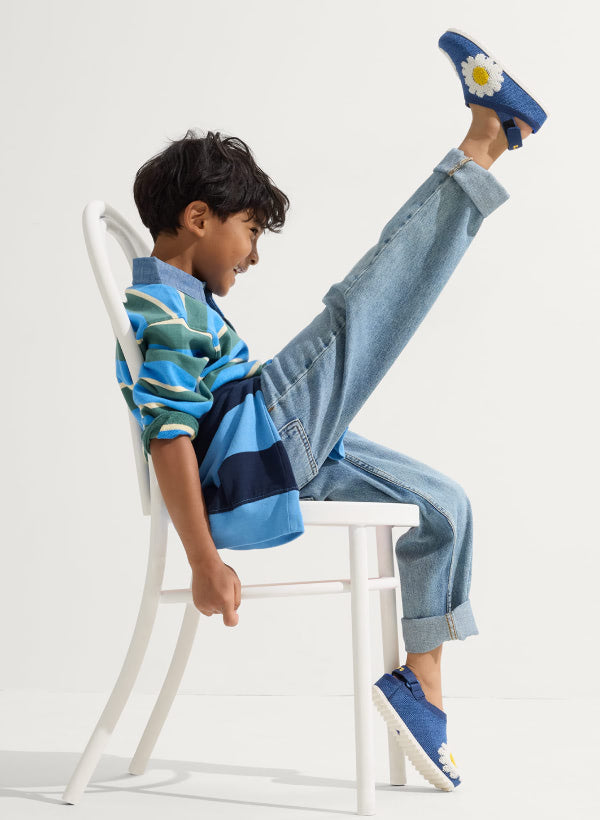 Child sitting on a white chair wearing The Kids Clog in Indigo Daisy, against a plain background.