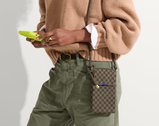 A woman in a sweater and pants holds a phone, featuring  the phone crossbody bag in canopy green