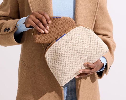 A woman in a tan coat holds two purses from the basketweave collection in vanilla wafer, showcasing stylish vanity set pouches.