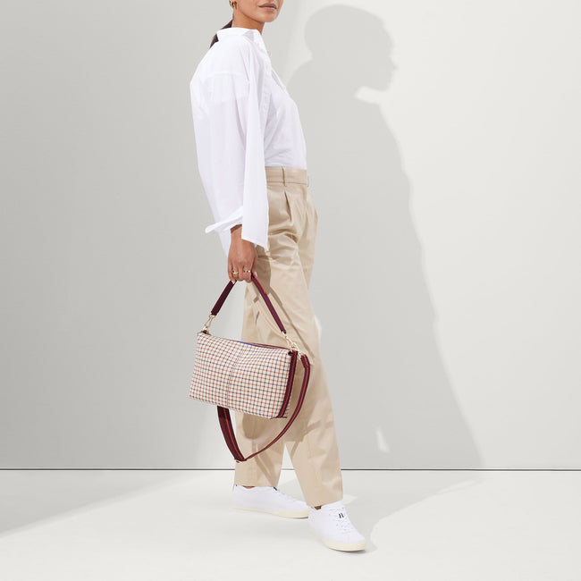 The Daily Crossbody in Malbec Grid, carried by its top handle by a female model, shown from the front.