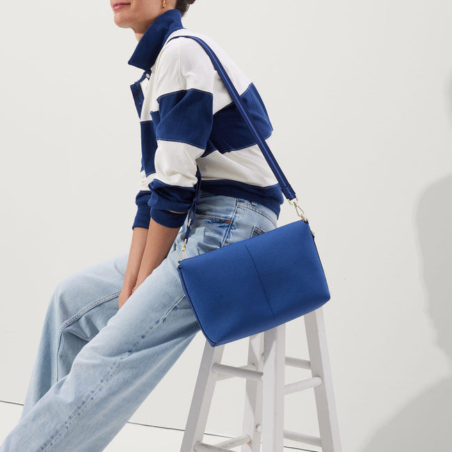 The Daily Crossbody in Varsity Blue, carried by its top handle by a female model, shown in motion from the right.