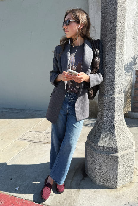 Woman leaning against a stone column on a sidewalk, wearing The Revelvet Penny Loafer in Revelvet Syrah." 