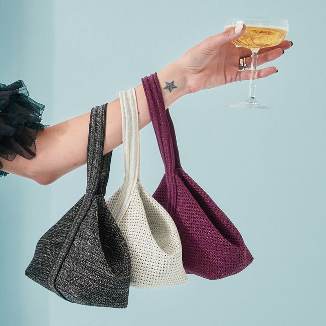 A model holding a glass of champagne and three different colors of The Party Pouch draped over her wrist: Onyx Sparkle, Diamond Sparkle, and Garnet Sparkle.