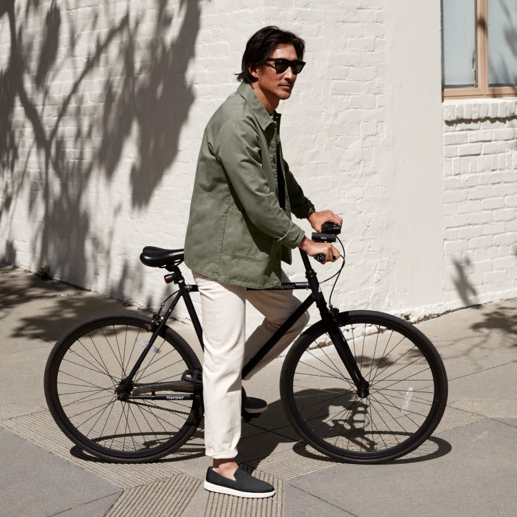 A model wearing The Ravello Slip On Sneaker in Black, riding a bike.
