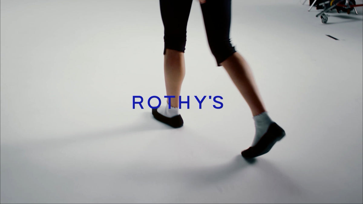 A person wearing black capri leggings, white socks, and black shoes walks across a light gray studio floor, with the blue text 'ROTHY'S' overlaid in the center.