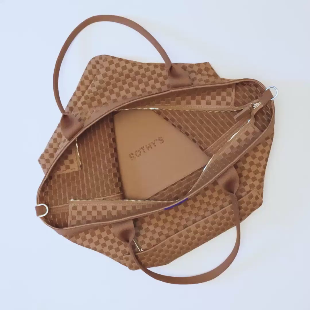 The Zip Travel Tote in Hazelnut shown being packed with a denim jacket, a sweater, a latop, a Vanity Set, a water bottle and a baseball cap. The model zips the bag closed and opens the small outer pocket, and places a pair of keys, a phone and a pair of sunglasses inside. The small pocket is zipped closed, and the model carries The Zip Travel Tote away by its top handles.