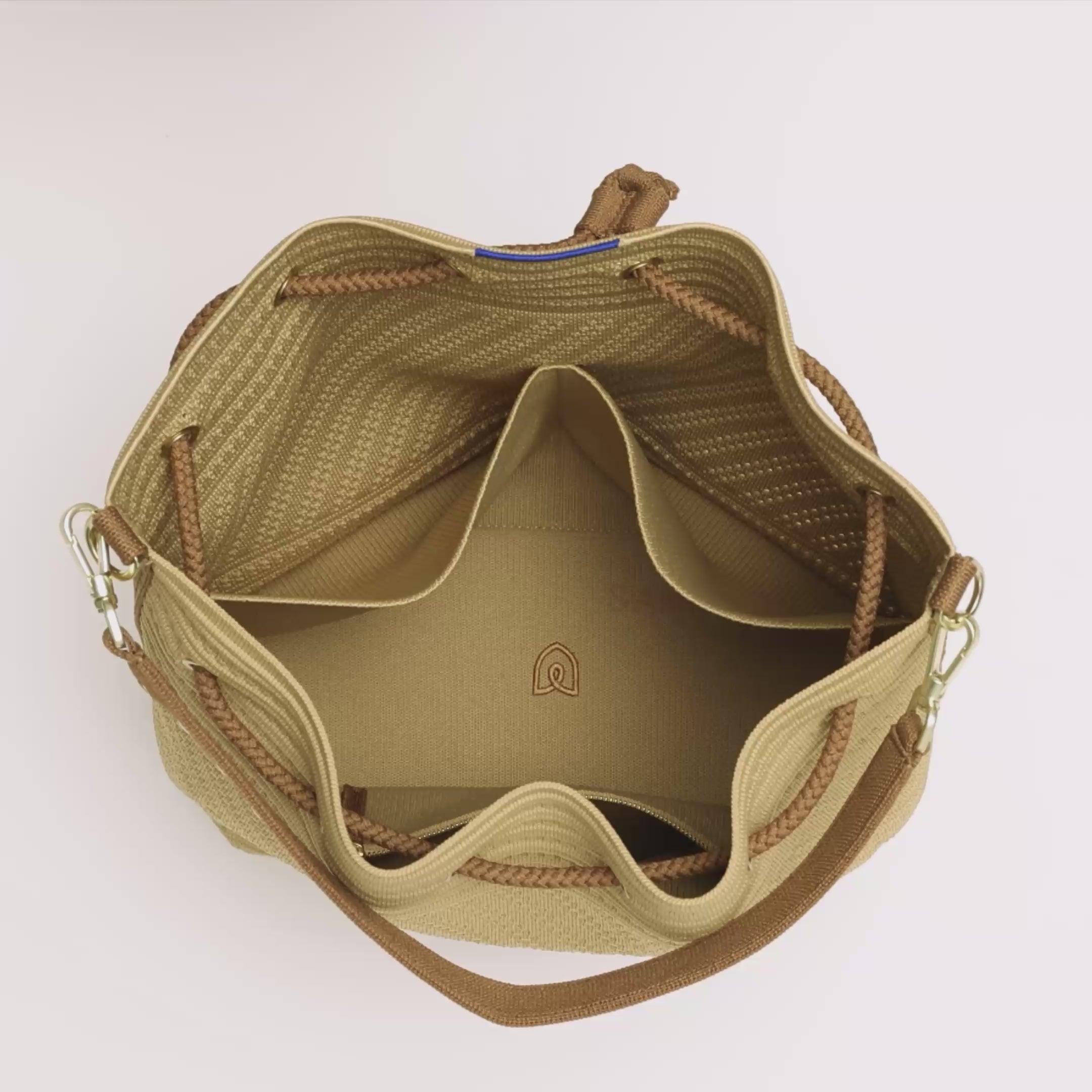 The Cinch Bucket Bag in Brown Butter Crochet is packed with a notebook, an iPhone, a laptop, water bottle, and travel pouch in the main compartment. In the interior pocket, the model packs smaller items before zipping it shut. They pull the main drawstring closure shut, grab the top handle and carry the bag out of frame.