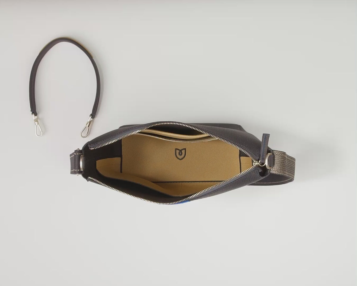 The Mini Zip Bucket in Ink & Ivory is shown unzipped from the top down, with its detachable top handle lying on its left side. A female model places in the main compartment a large notebook and a medium-sized notebook, a water bottle and the Mini Universal Pouch. She then places in the interior slip compartments a tin of Altoids, an energy bar, sunglasses and lip balm. Then she places her iPhone and airPods in the interior zip pocket. 