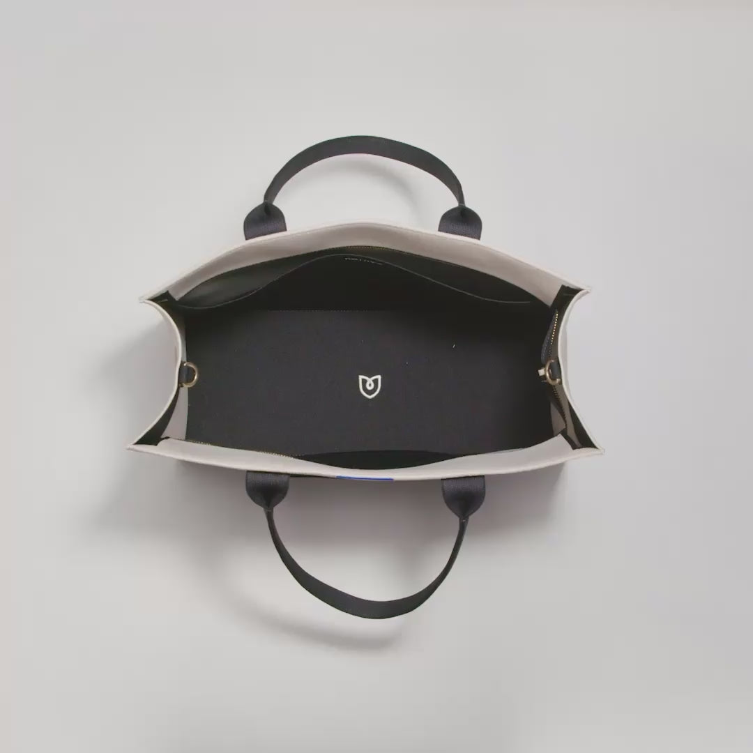 The Classic Tote in Luna shown packed with a laptop, a notebook, The Vanity Set in Black Mist, a book, a notepad and pens, a water bottle, airpods, cleaning wipes and keys. A female model then zips up the bag, unzips the exterior zip pocket and packs an iPhone. The model then zips it back up and carries the bag out of frame.