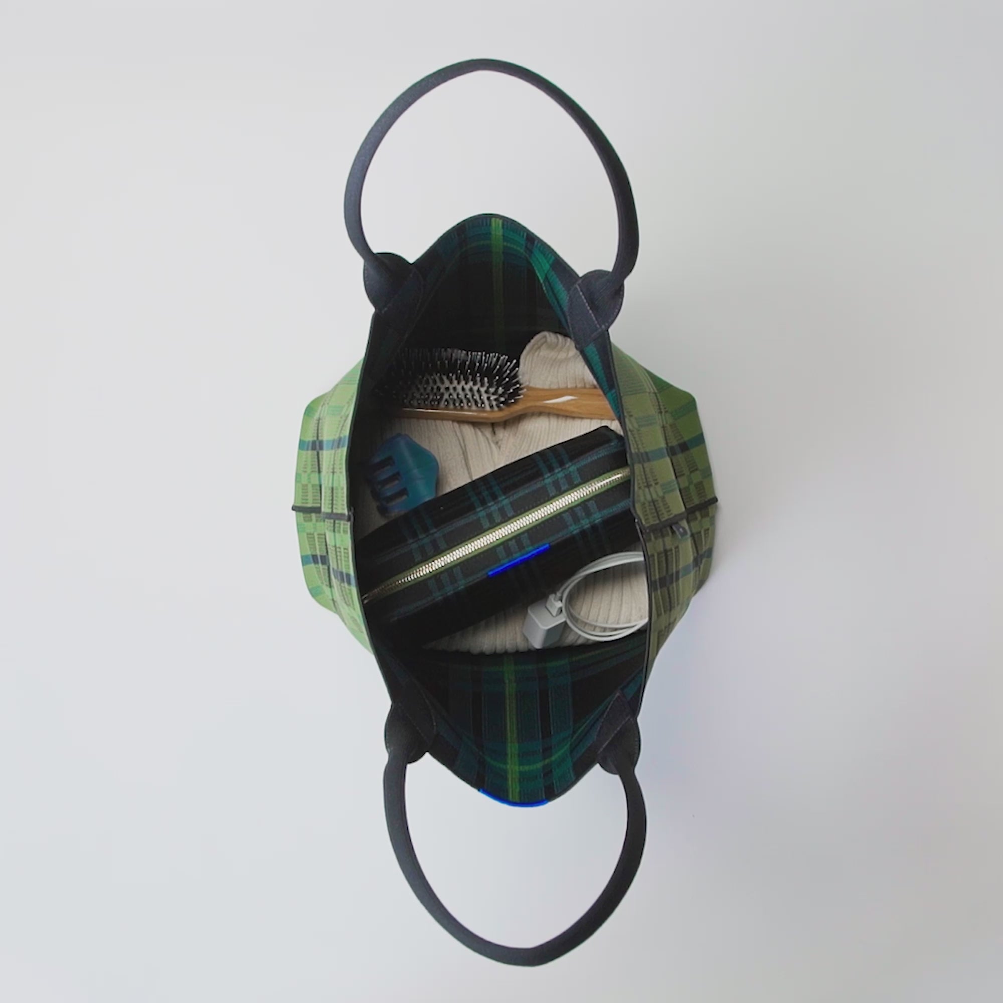The Reversible Lightweight Tote in Pine Tree Plaid shown packed with The Universal Pouch in Cedar Plaid, a hair clip, a charger, and hair brush, a sweater and a book. Model unpacks listed items, turns The Reversible Lightweight Tote inside out, and packs everything back again. Model then loops a key leash around one of the tote straps, clips The Mini Universal Pouch to the key leash, picks up the handles and carries the tote out of frame.