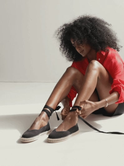 A model wearing The Espadrille in Black, tying the removable ankle straps.