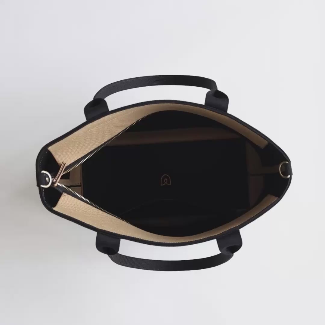 The Lightweight Zip Tote in Black Sky being packed with a laptop, notebook, Wallet Wristlet, water bottle, apple, hand cream, a pair of keys, an iPhone, a keyring and a pair of sunglasses. The model zips the bag and carries it away.